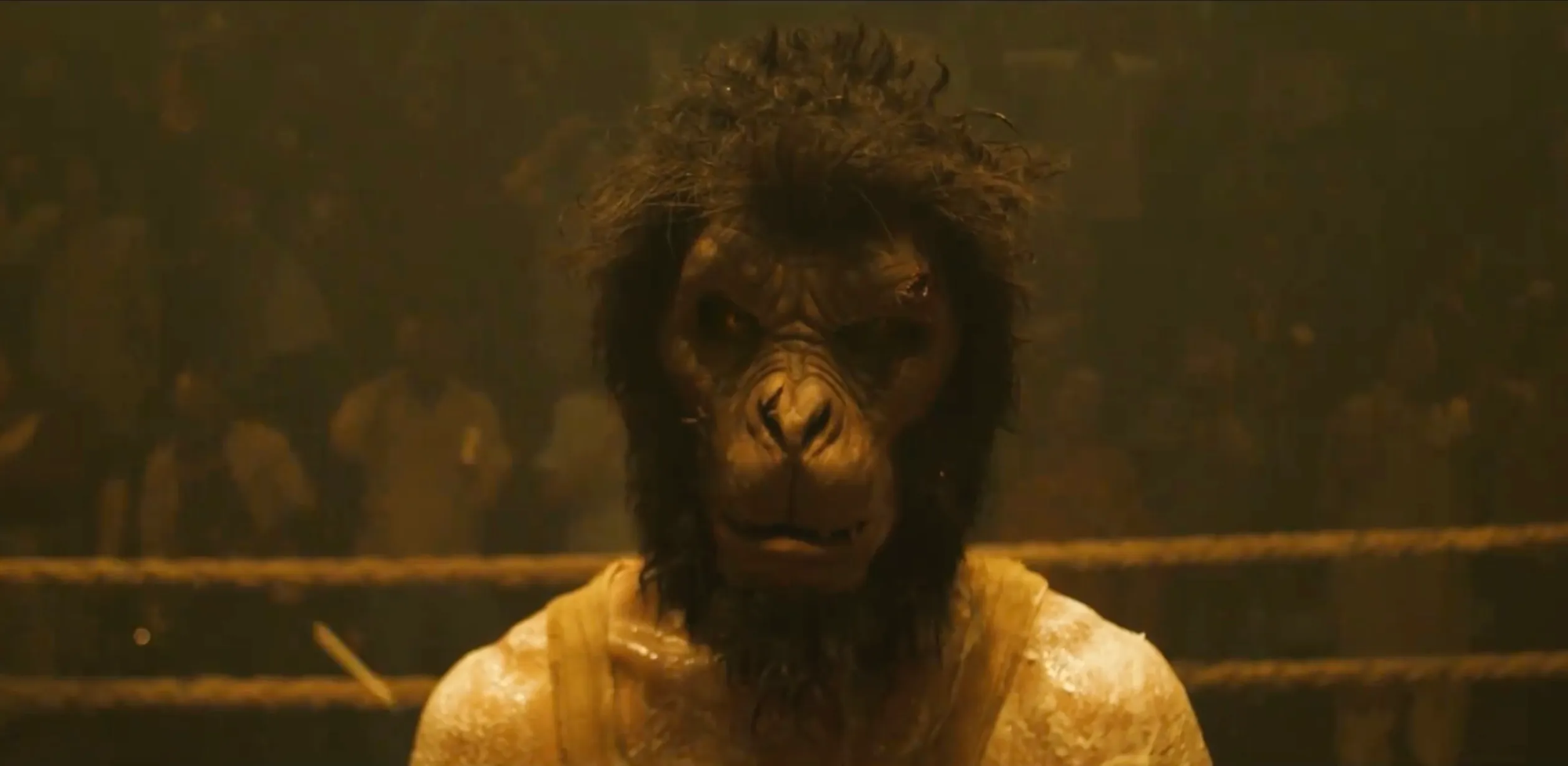 A shot from Monkey Man. The main character stands in a fighting ring wearing a terrifying and filthy monkey mask. His shoulders are covered in scars. A chanting crowd is visible behind him.