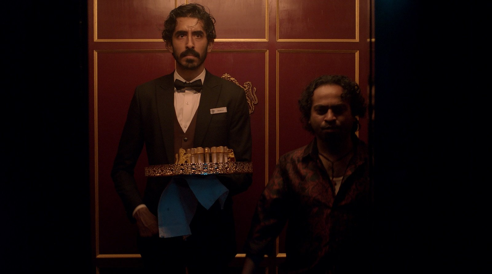 A shot from Monkey Man. The main character (The Kid) and his workmate arriving at a party in a fancy elevator. The Kid is holding a tray of party favours for guests and is dressed in a waiter's uniform with a bow tie. The elevator is lined with red panels.