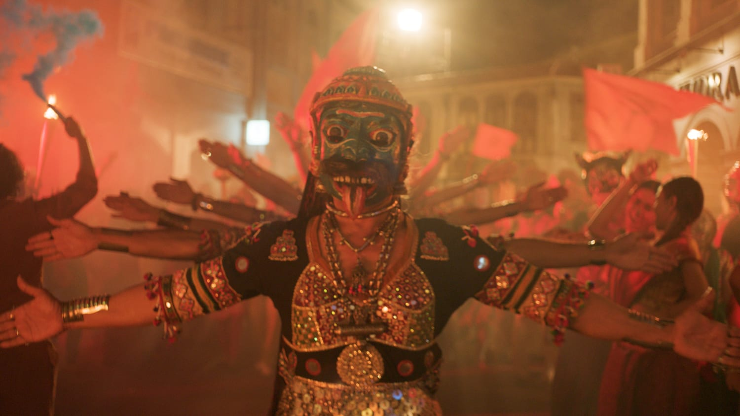 A shot from Monkey Man. Hijra dancers spreading their arms in the street. The front dancer is wearing a blue mask with a tongue poking out. There are celebrations happening around the rest of the street, with flags and flares.